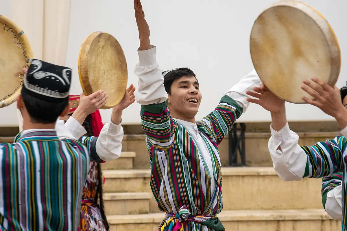 Male dancers in striped robes and embroidered skullcaps raise doira frame drums during an Uzbek classical dance ensemble performance