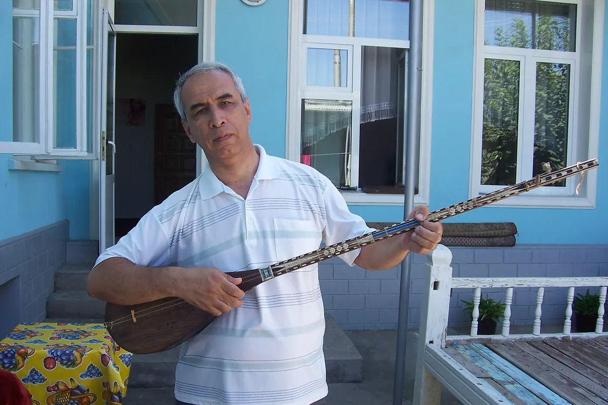 A man holds a dutor, a long-necked two-string lute with an inlaid neck, in front of a blue house