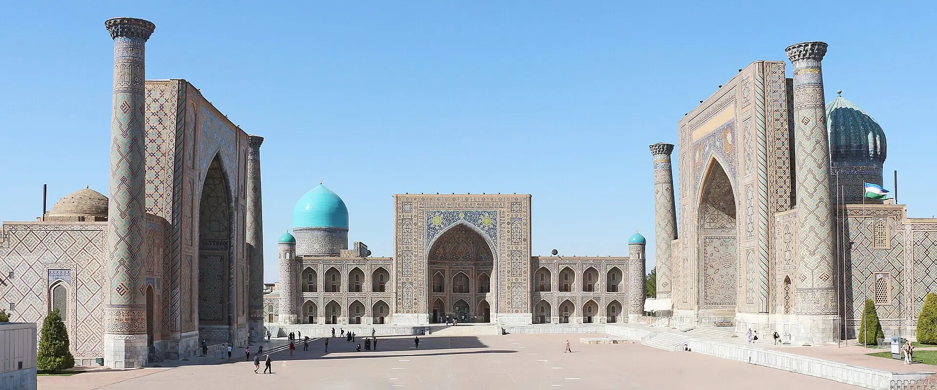 Registan Square in Samarkand under a clear blue sky, with three madrasas around the square, tall tiled minarets and turquoise domes