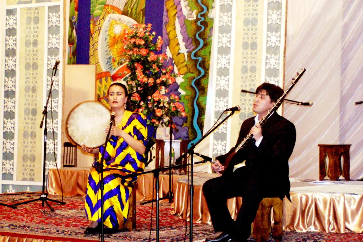 A woman in a yellow and blue ikat dress plays a doira frame drum beside a man playing a long-necked lute on a stage with carpets and flowers