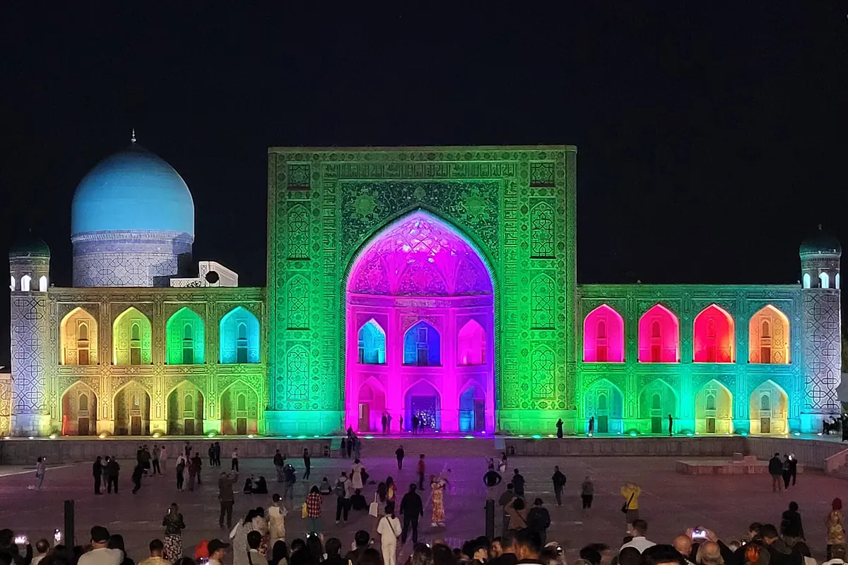 The Tilya-Kori Madrasa on Registan Square in Samarkand lit in green, magenta and gold at night, with a crowd in the square